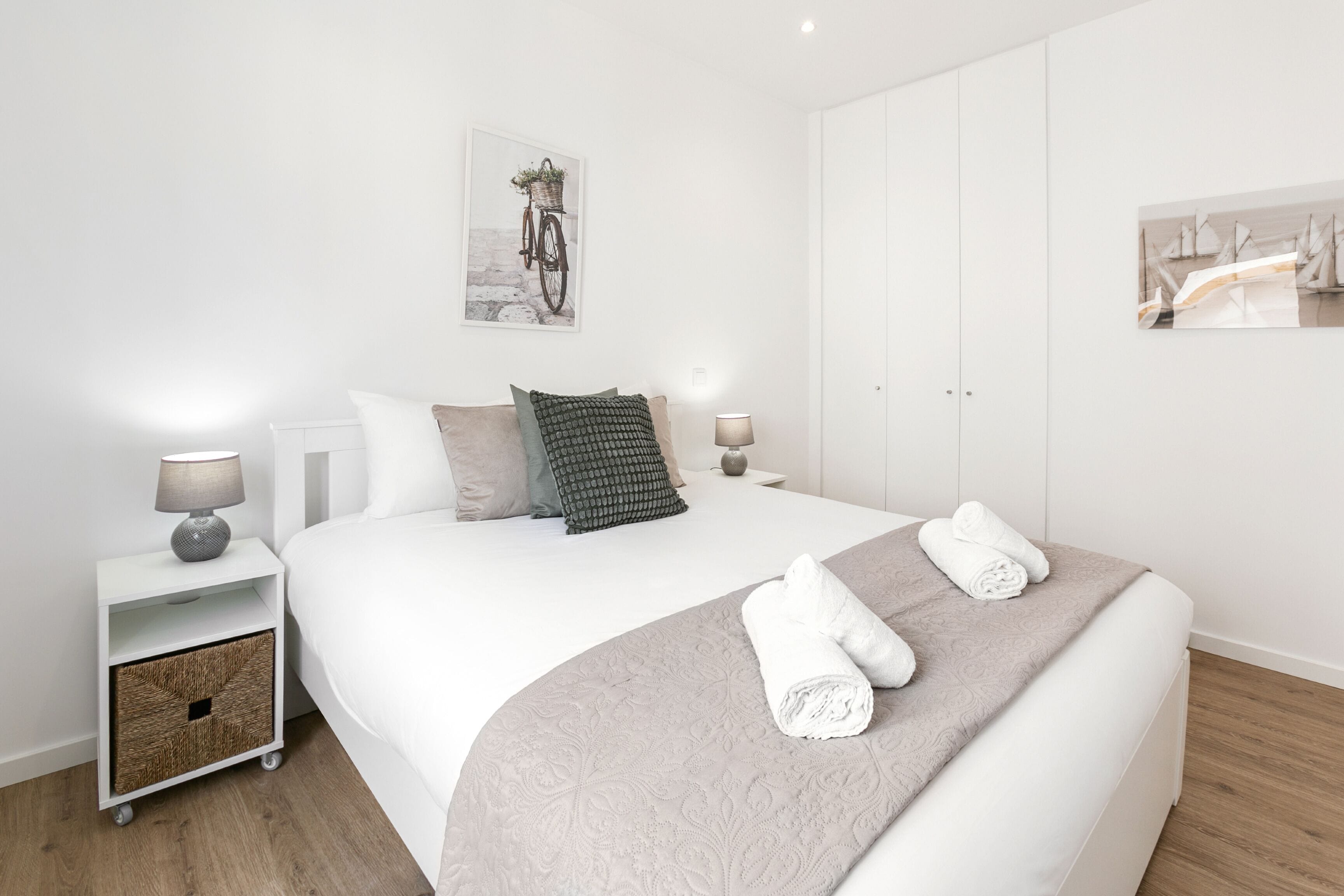 A neatly arranged bedroom with a large bed, two bedside tables with lamps, a small storage unit, and artwork on the walls.