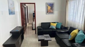 Una sala de estar con un sofá negro, una mesa de centro y un mueble para televisión. Se ve una puerta que da a otra habitación y un cuadro en la pared.