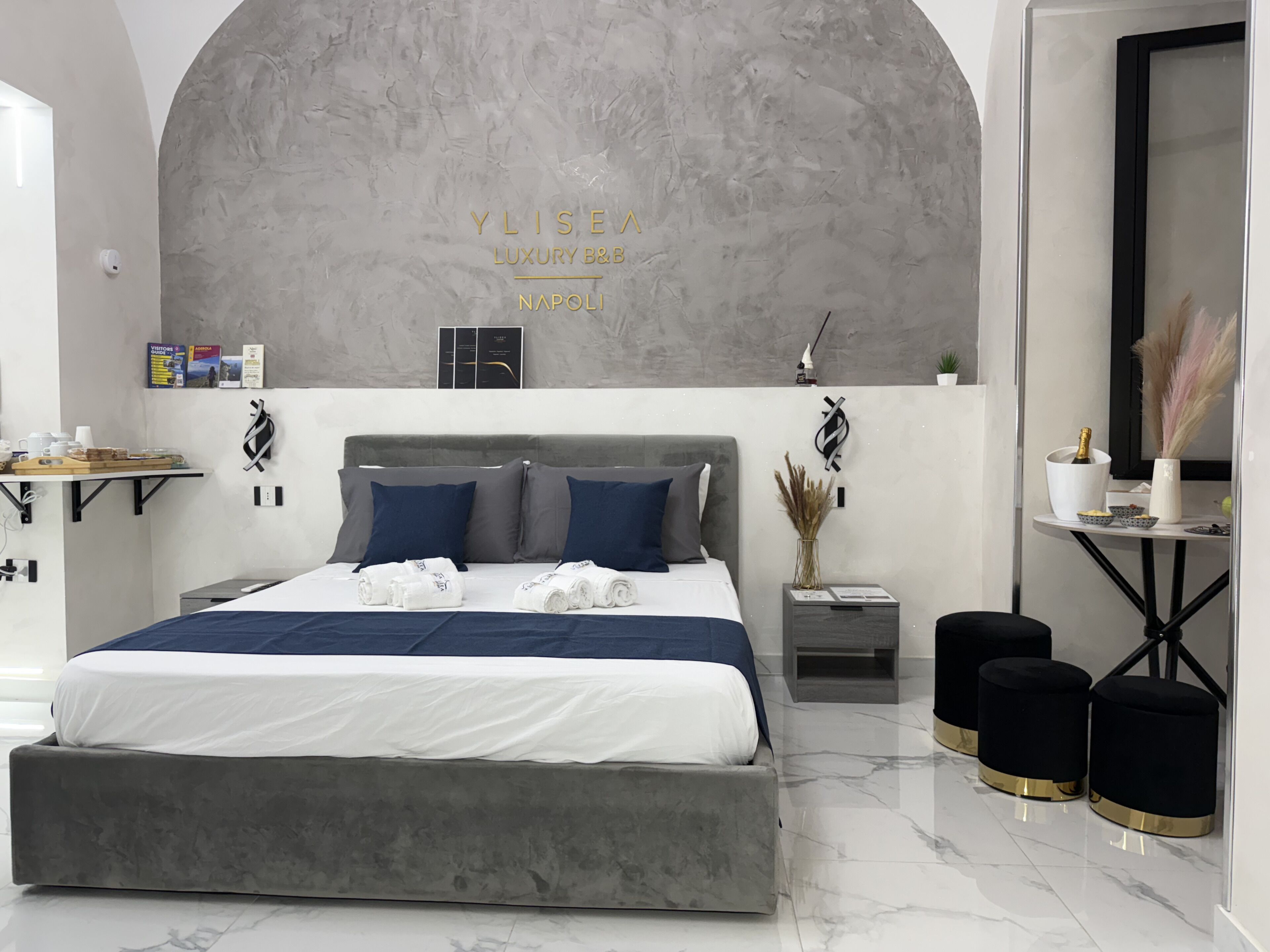 A modern bedroom with a large bed, a grey headboard, and a wall-mounted artwork reading "YLISEA LUXURY B&B NAPOLI".