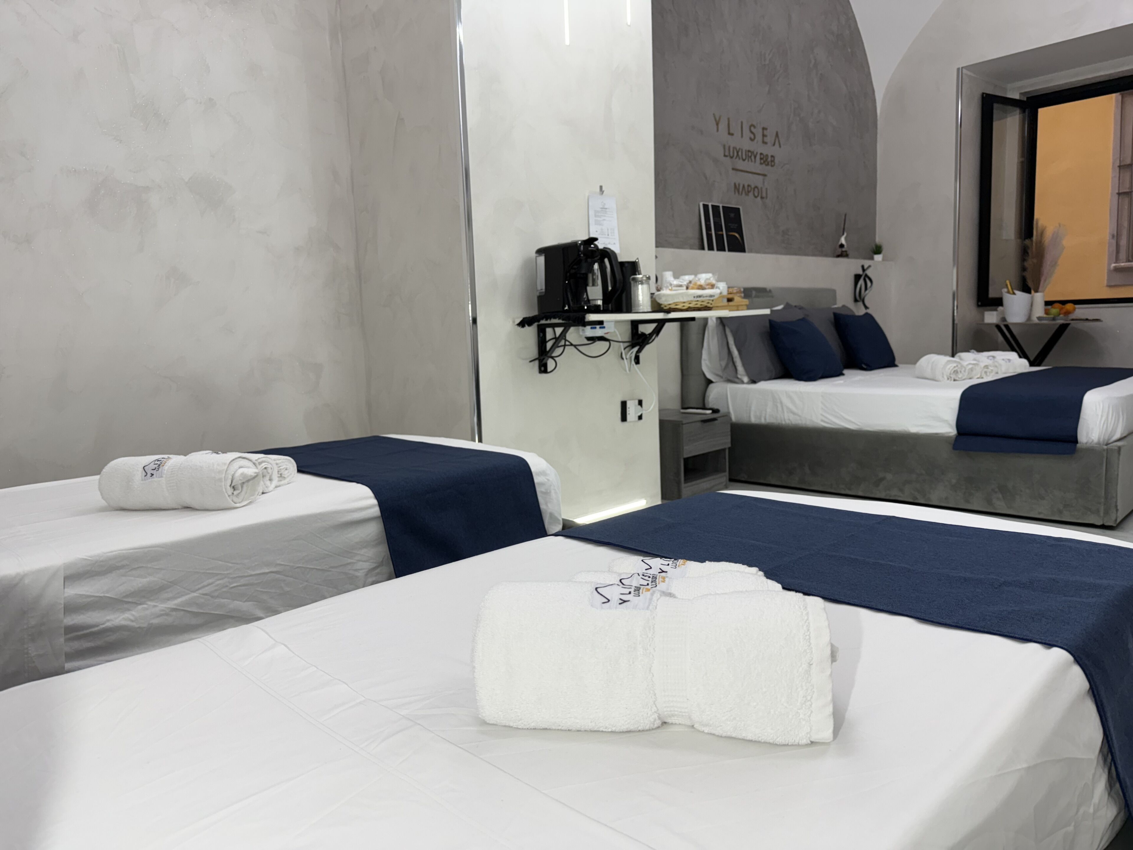 A hotel room with two beds, each with a blue bedspread and white linens. There is a small table with a coffee machine and a basket of fruit. The room has a modern design with a grey wall and a sign that reads "YLISEA LUXURY B&B NAPOLI".
