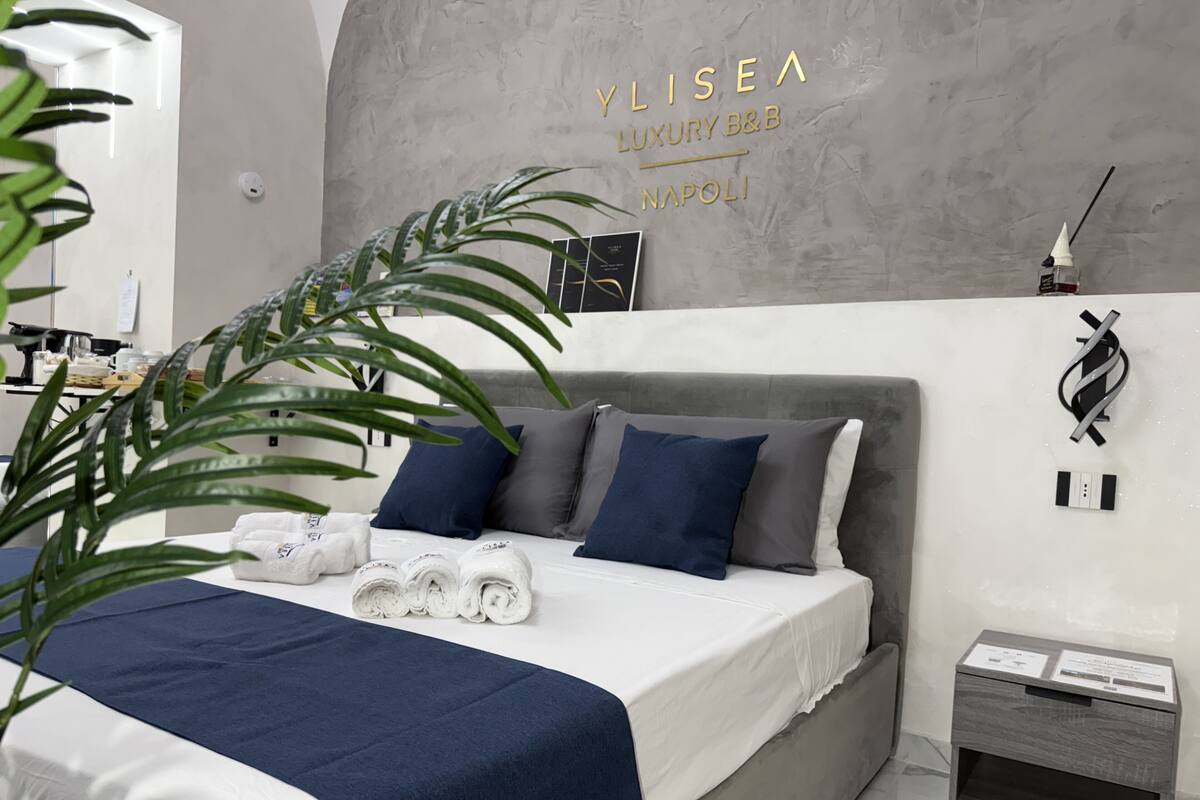 A modern bedroom with a grey headboard, white bedding, and blue pillows. There are rolled towels on the bed and a small bedside table with a lamp.