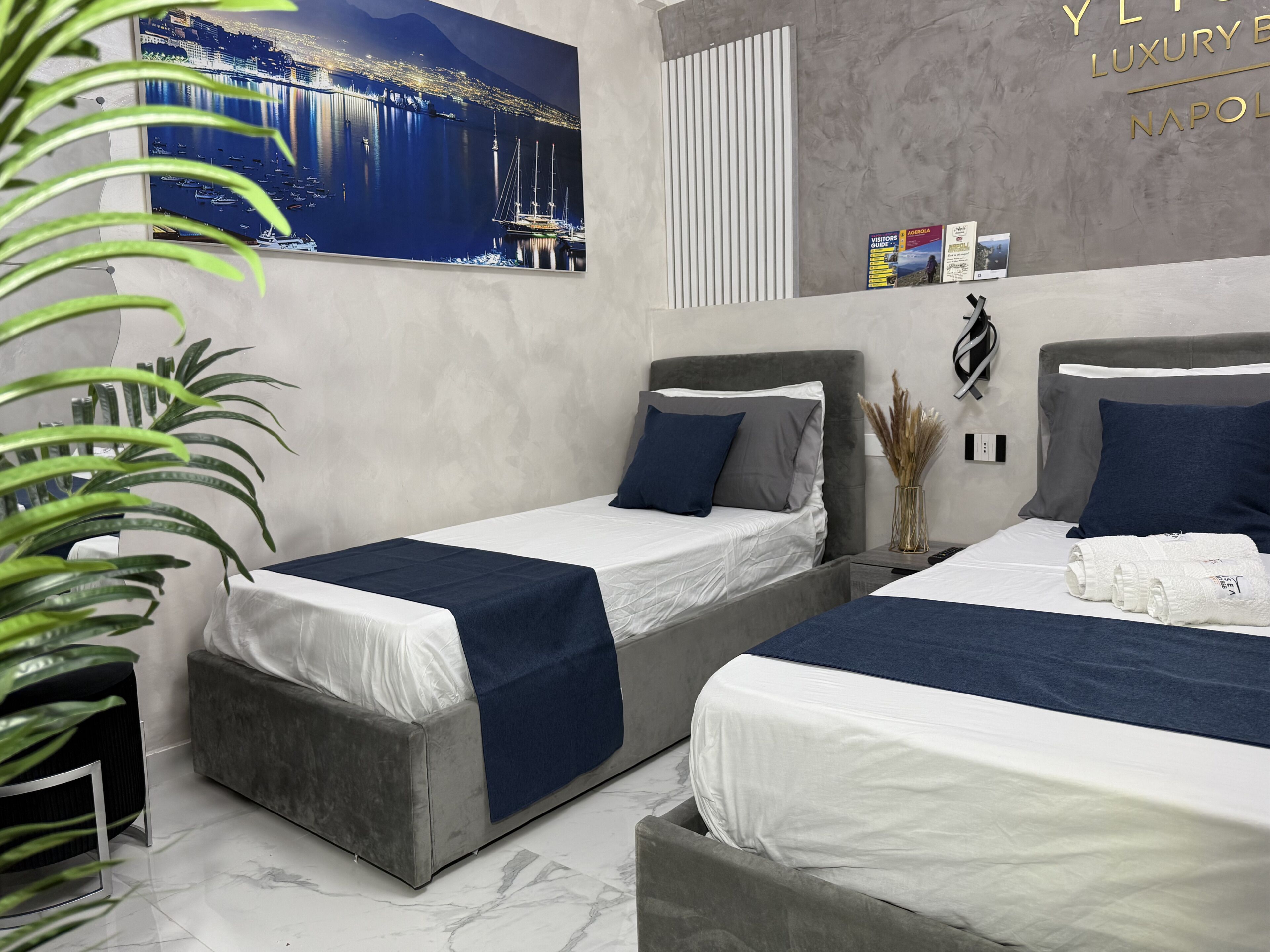 A hotel room with two beds, a large cityscape painting, and a wall-mounted sign reading "LUXURY NAPOLETANO".