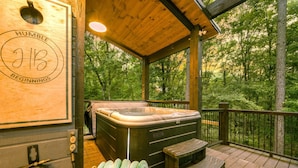 Eine Holzdar Terrasse mit Whirlpool, eine Holzverkleidung mit einem Logo und Blick auf einen Wald.