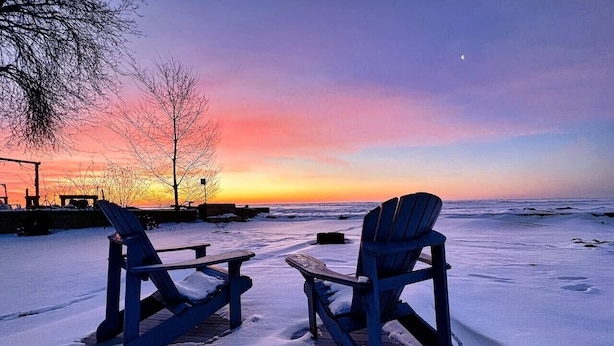 Dos sillas Adirondack y una mesa de picnic sobre una playa nevada con un atardecer de fondo.