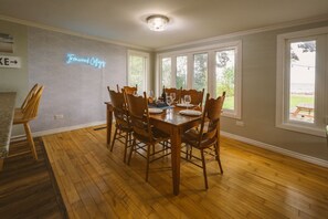 A dining area with a wooden table and chairs, a neon sign reading "Trinity Cottages", and large windows offering a view of the outdoors. - Sauna, stunning views of Lake Erie, lakefront retreat (Kingsville)