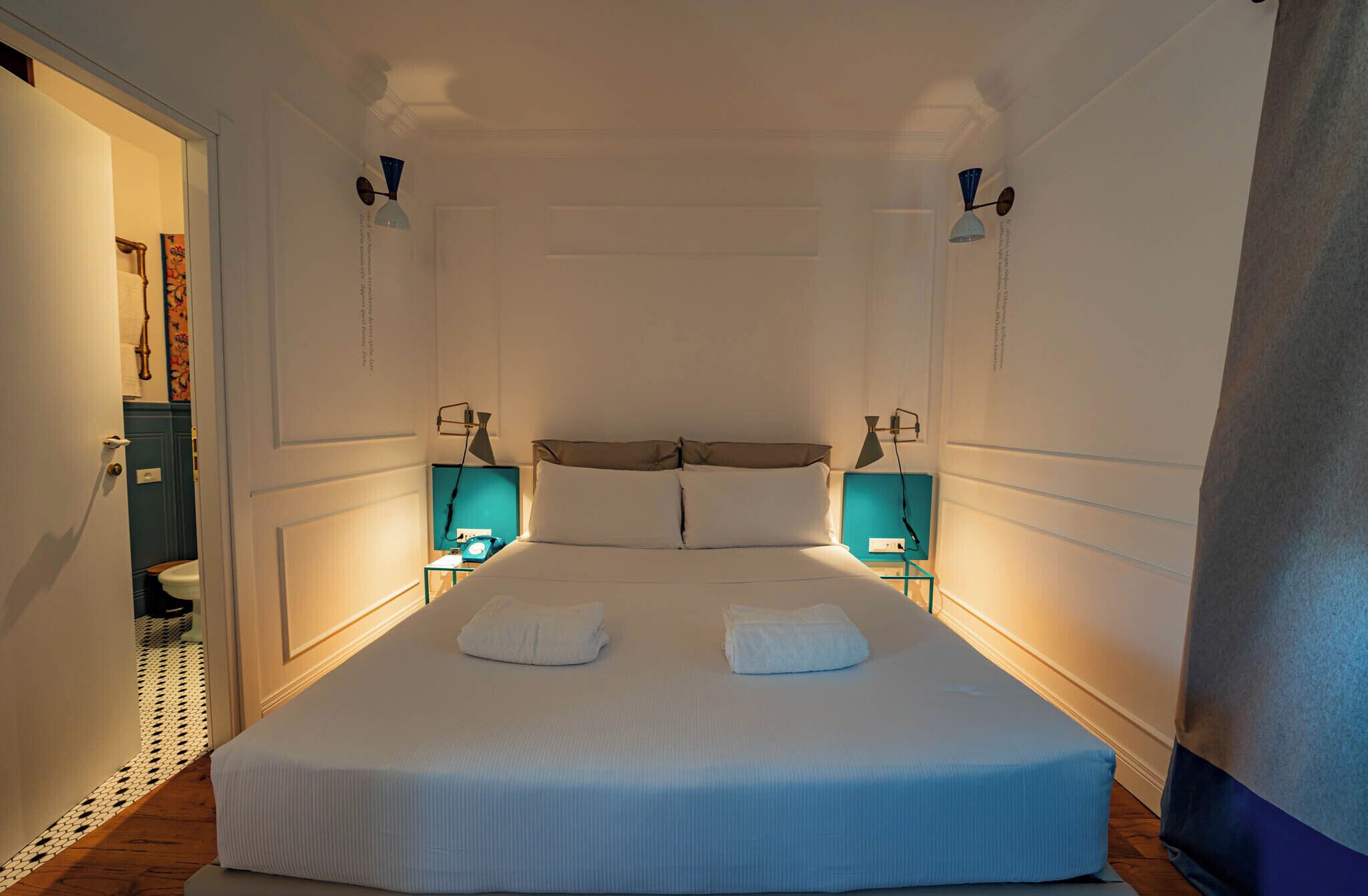 Un dormitorio con una cama grande, dos mesitas de noche con lámparas y vista al baño a través de una puerta abierta.