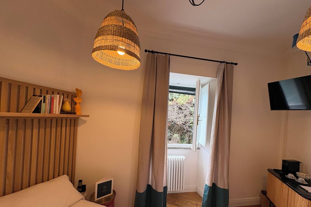 Un dormitorio con una cama, una mesita de noche, una estantería con libros y un florero, una ventana con cortinas y un televisor montado en la pared.