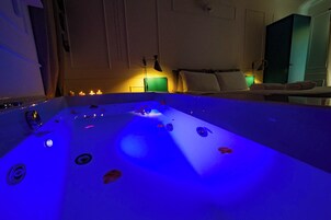 Una habitación con una bañera grande, una cama con almohadas, una mesita de noche con lámpara y un armario.