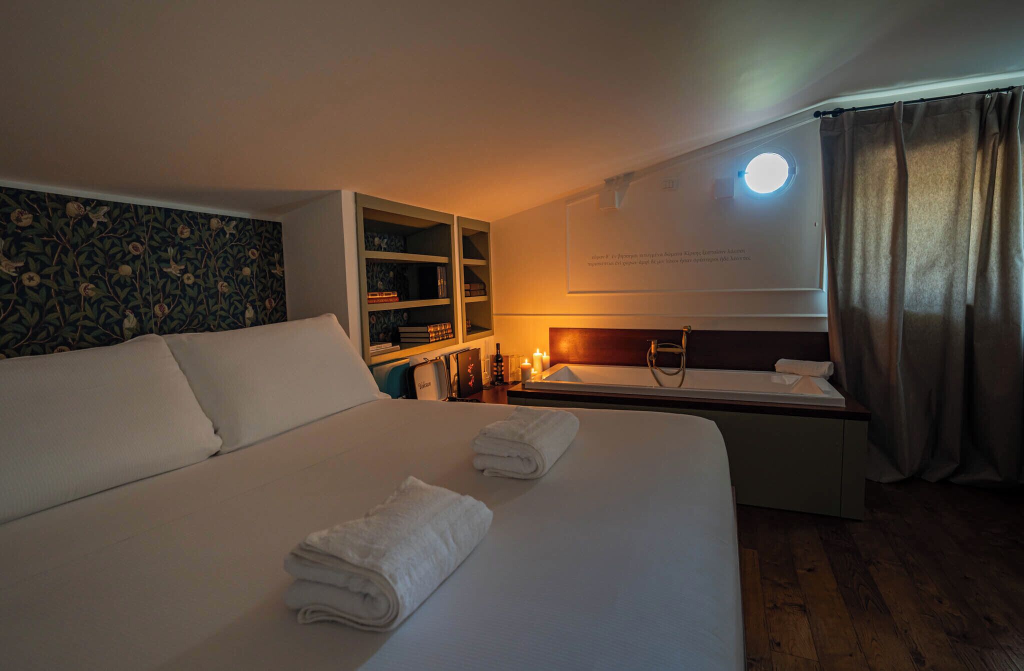 A hotel room with a large bed, a bedside table with a lamp, a small shelf with books, and a window with curtains.