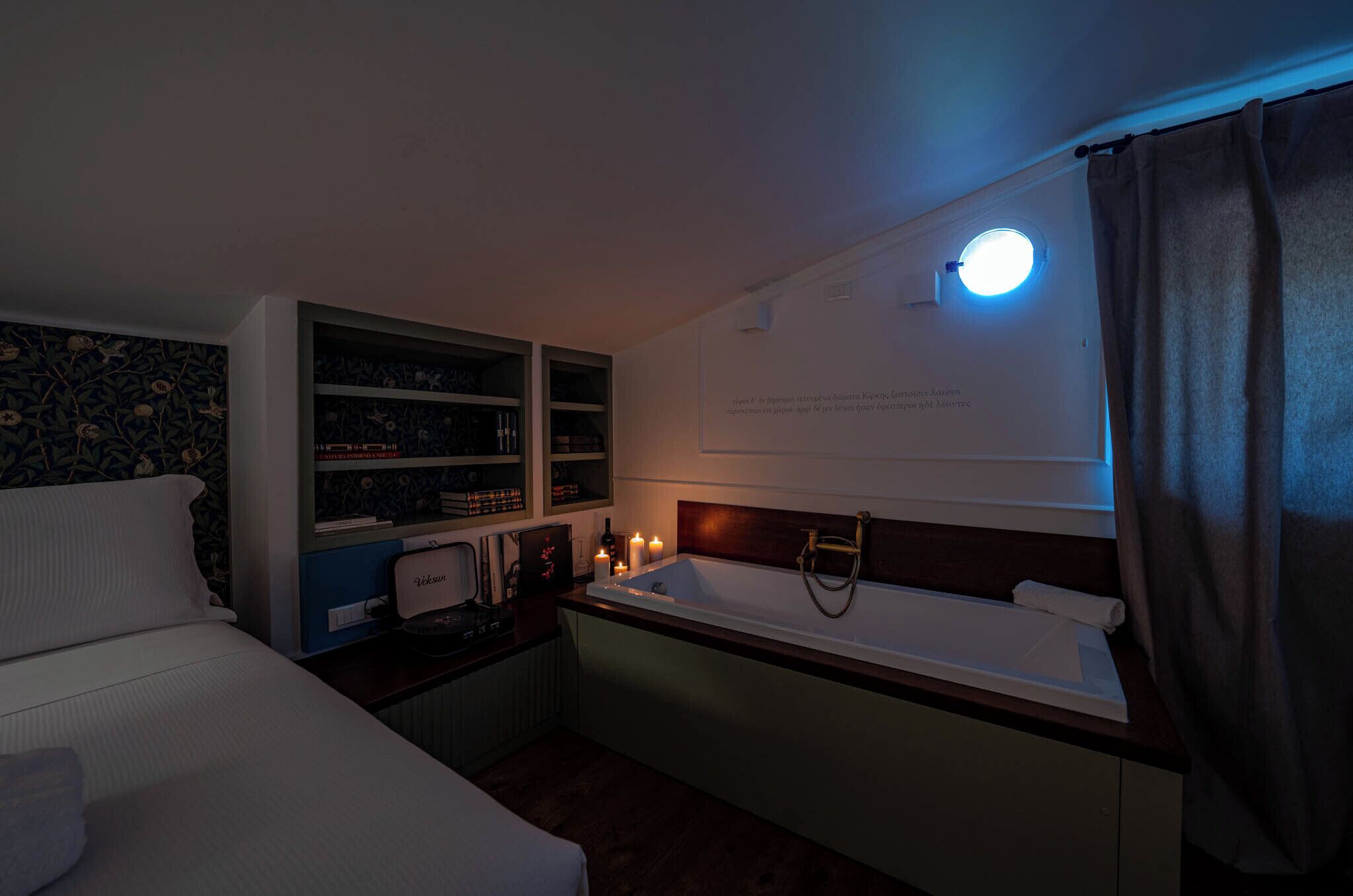 A hotel room with a bed, a small desk with a lamp, a vanity with a mirror, a bathtub, and a window with a curtain.