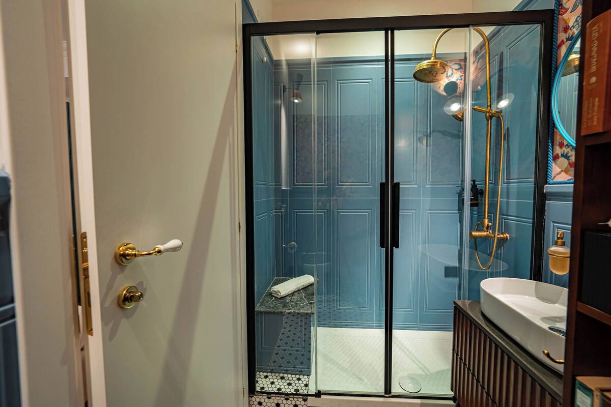 Un baño con puerta de vidrio, ducha con cabezal dorado y lavamanos blanco.