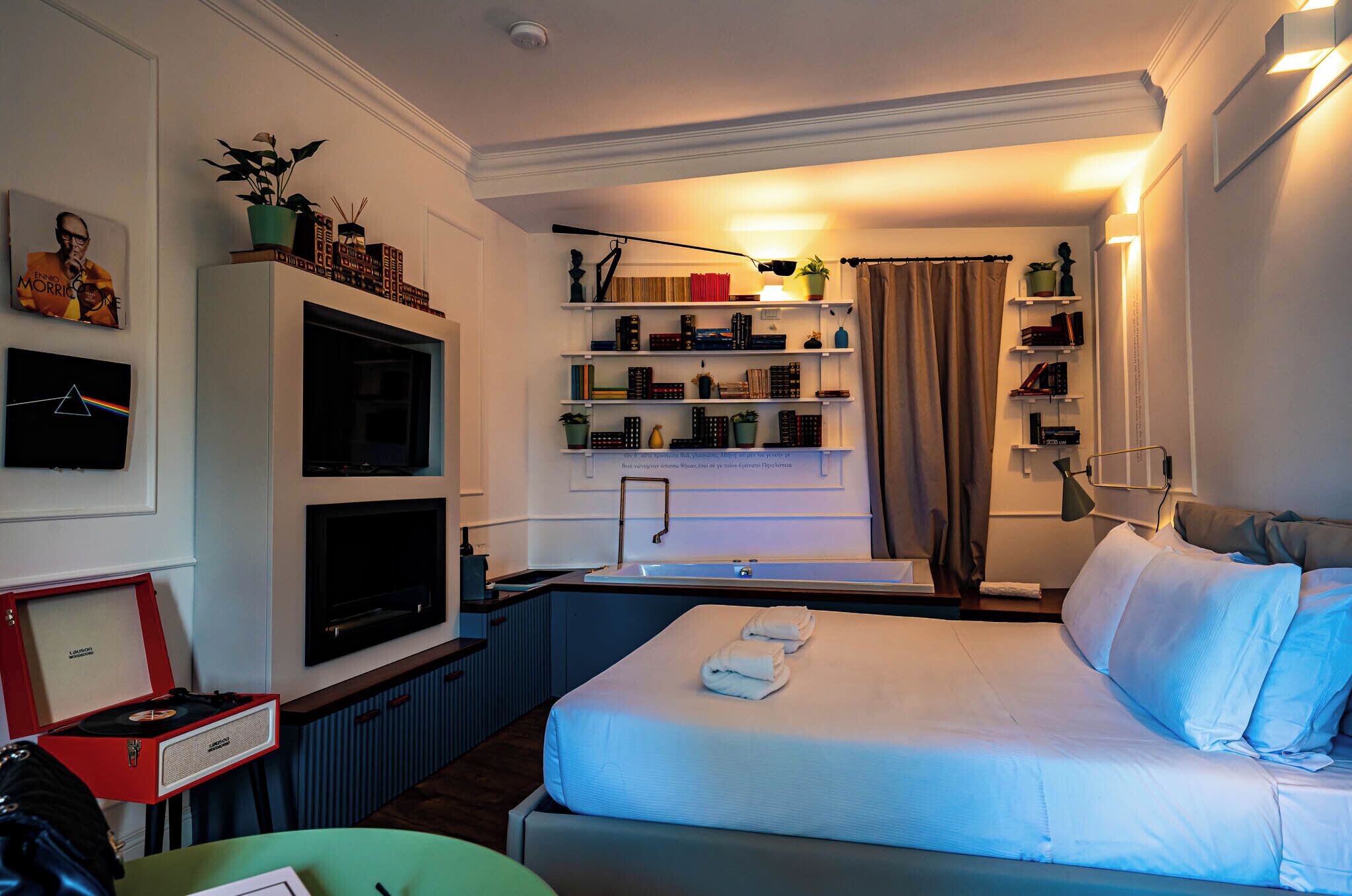 A hotel room with a bed, a TV, a bookshelf, and a desk with a red speaker.