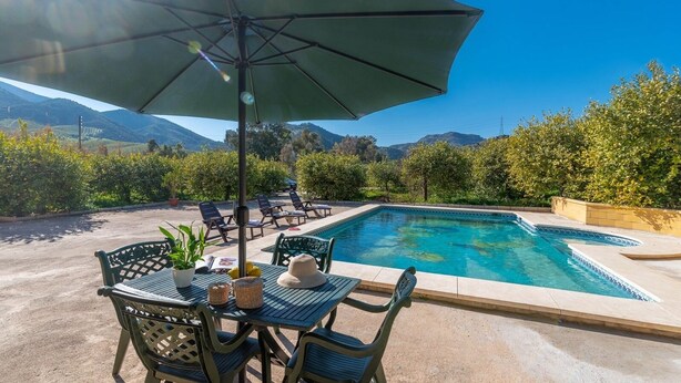 Poolbereich mit Liegestühlen und einem Tisch, umgeben von Grünflächen und Bergen im Hintergrund.