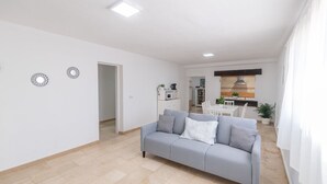 Ein modernes Wohnzimmer mit einer grauen Couch, einem Essbereich mit weißem Tisch und Stühlen sowie einer Küche im Hintergrund.