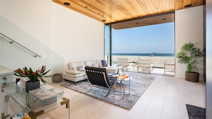 Un soggiorno moderno con una parete vetrata che si affaccia su una spiaggia, un soffitto in legno e una scala in vetro.