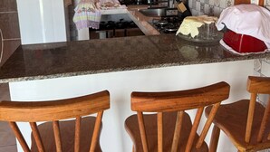 A kitchen with a granite countertop, a gas stove, a coffee maker, and a refrigerator. There are three wooden chairs in front of the counter. - Chalé panulirus (Icapuí)
