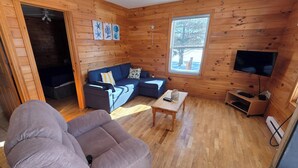 Une chambre confortable, aménagée dans le style d’un chalet, avec un intérieur en bois, un canapé, une petite table, un téléviseur et une fenêtre donnant sur la forêt.