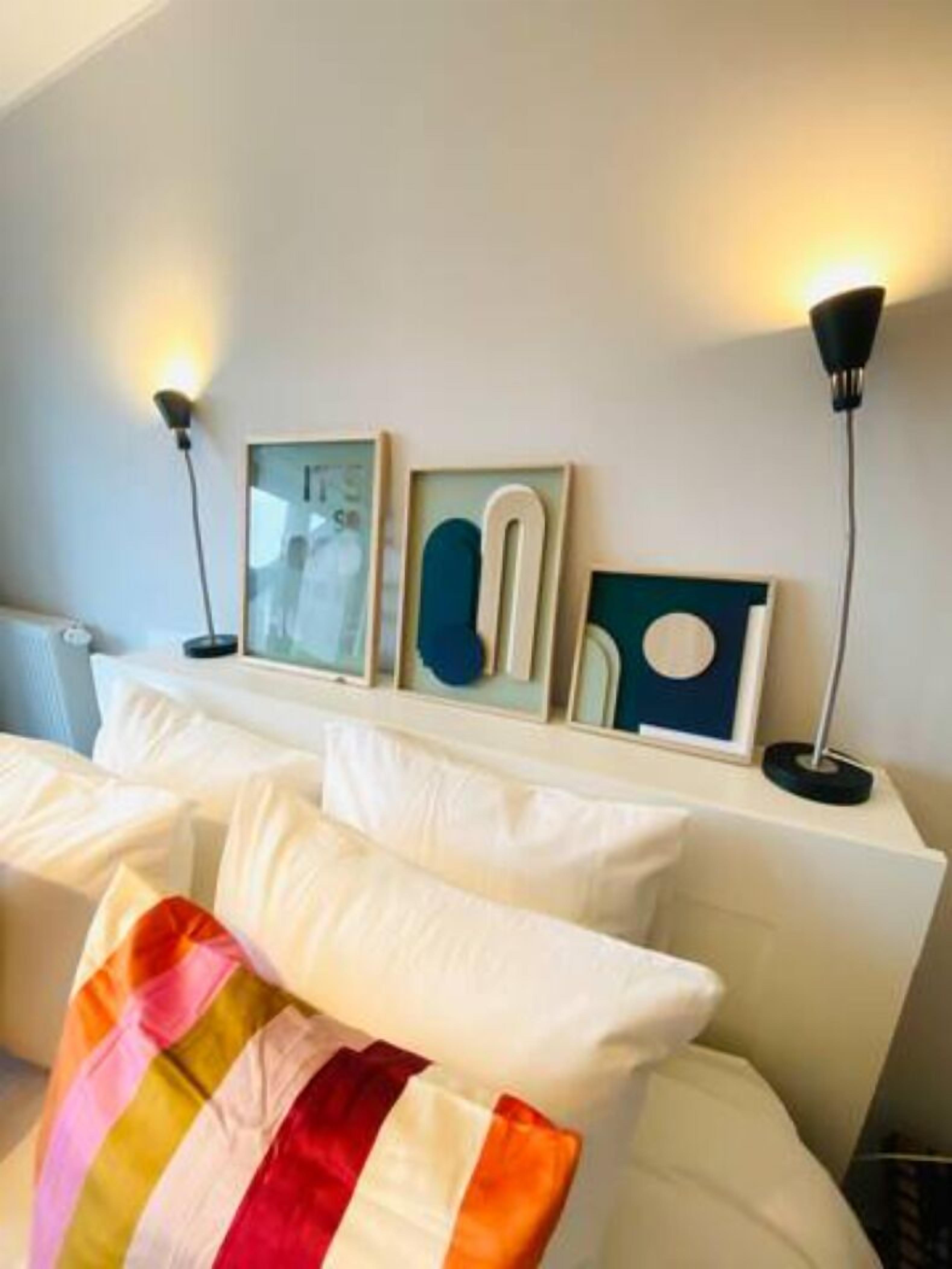 Une chambre d’hôtel avec un lit, du linge de lit blanc, un coussin rayé aux couleurs vives et des œuvres d’art accrochées au mur.