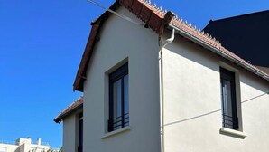 Edificio residencial de dos pisos con techo de tejas, cerca de madera y un pequeño cuadro eléctrico en la planta baja.