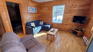 Ein gemütliches Blockhauszimmer mit Holzvertäfelung, einer Couch, einem kleinen Tisch, einem Fernseher und einem Fenster mit Blick auf Bäume.