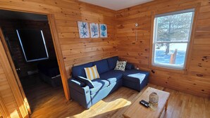 Ein gemütliches Blockhaus-Interieur mit einer Eckcouch, einem kleinen Holztisch und einem Fenster mit Blick auf Bäume.