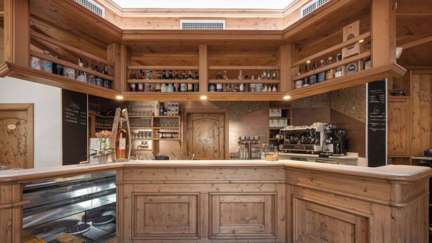 A wooden interior with a central counter, shelves with bottles, and a coffee machine. - Grünwald (Salorno)