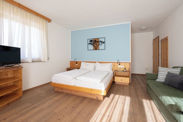 A bedroom with a wooden bed, a television on a wooden stand, a green sofa, and a painting on the wall. - Grünwald (Salorno)