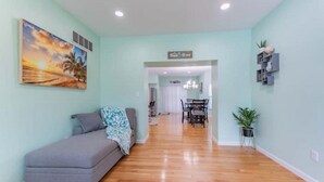 A living room with a grey sofa, a painting of a sunset, wooden flooring, a dining area with a chandelier, and a sign indicating a 'Living Room'. - Perfect Getaway @ CWE- Entire Home + Free Parking Near Fox, Universities & More! (St. Louis)