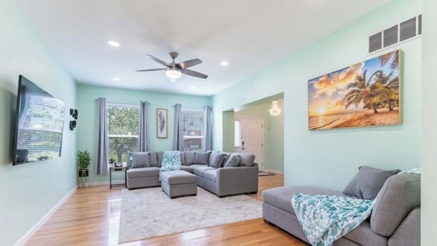A spacious living area with a large gray sectional sofa, a smaller gray sofa, and a coffee table. There is a ceiling fan, a painting of a beach scene, and a window with curtains. - Perfect Getaway @ CWE- Entire Home + Free Parking Near Fox, Universities & More! (St. Louis)