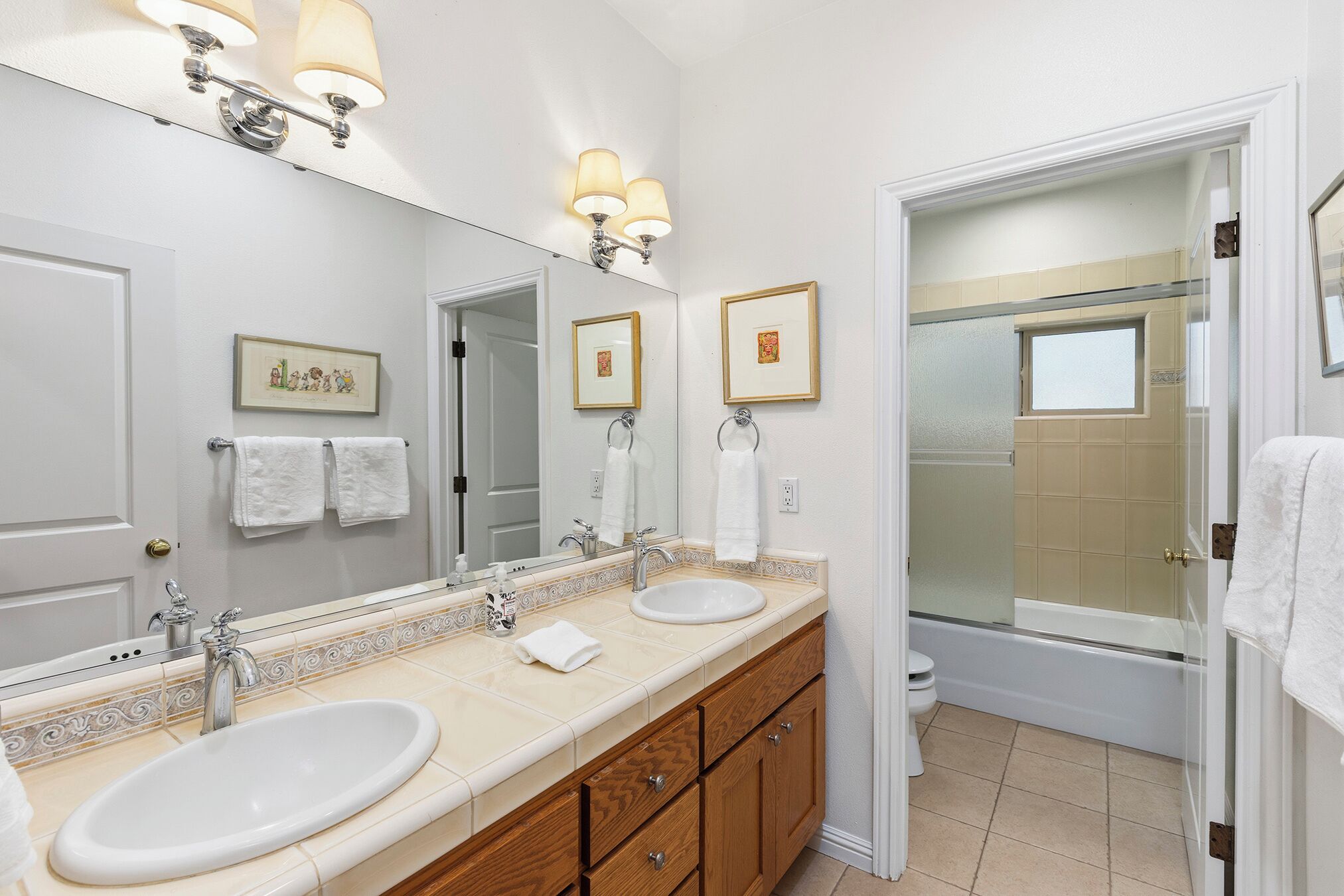 A bathroom with two sinks, a large mirror, wall-mounted lights, framed pictures, and a shower area.