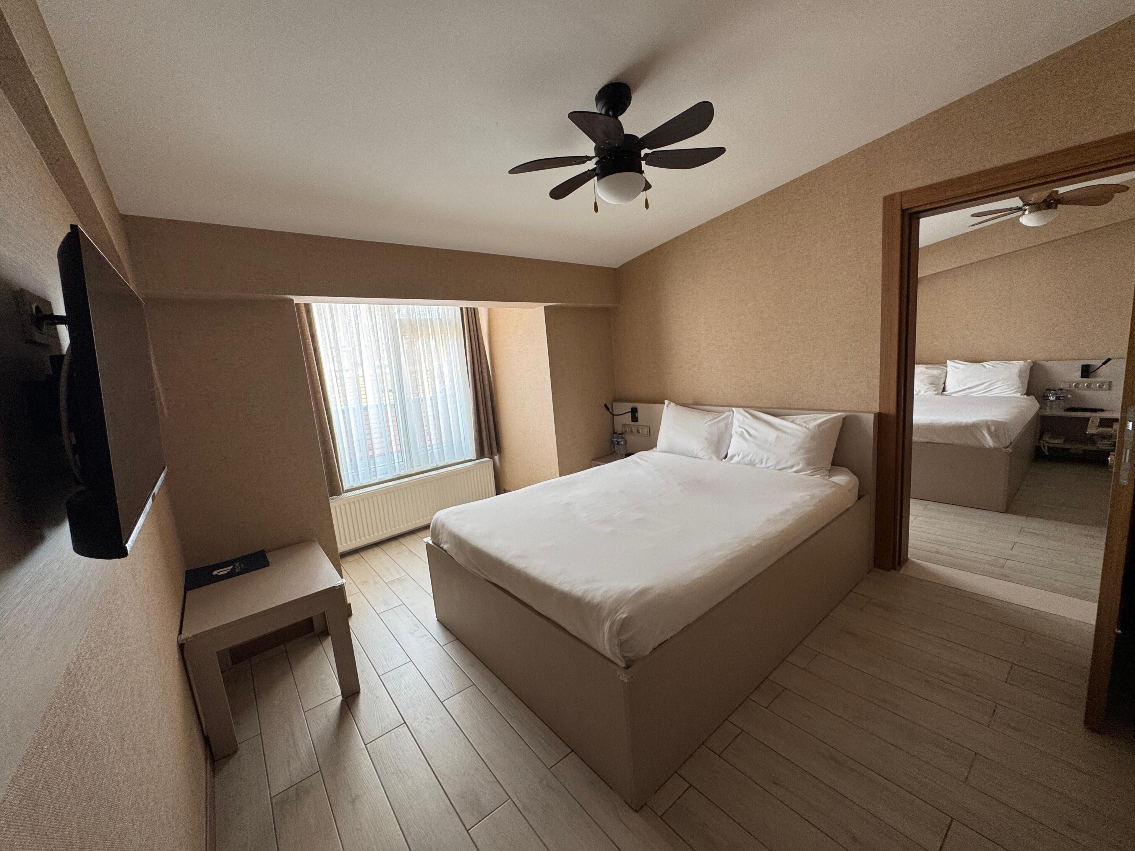 A hotel room with a bed, a ceiling fan, a window with curtains, a bedside table, and a mirror.