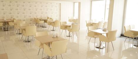 Un comedor amplio y luminoso con varias mesas y sillas, ventanas grandes y suelo de baldosas.