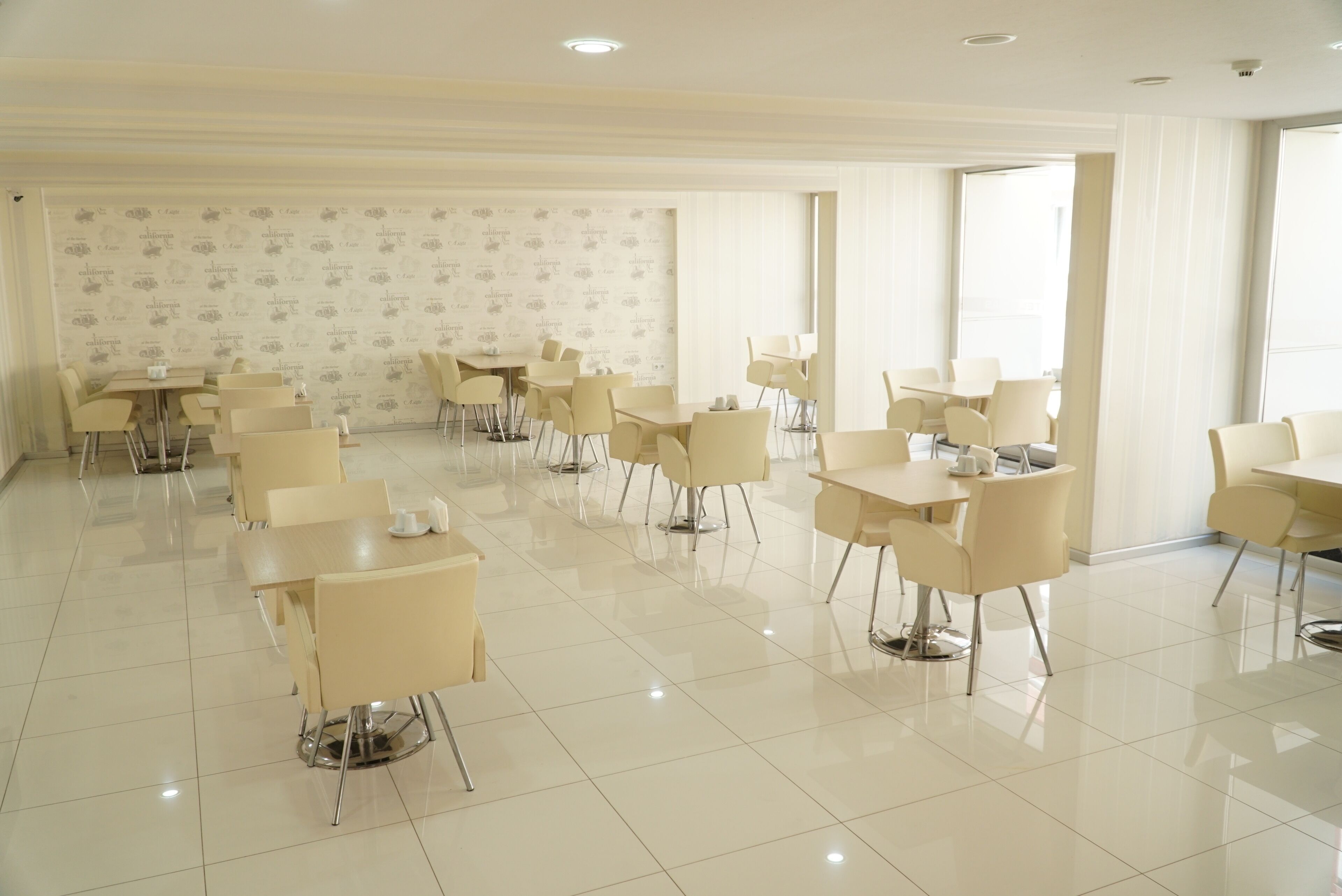 A spacious, well-lit dining area with modern, cream-colored chairs and tables, featuring a clean, tiled floor and a patterned wallpaper on the back wall.