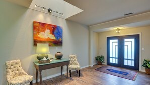 Un hall d’entrée spacieux avec un plancher en bois, deux fauteuils rembourrés, une table avec une lampe, un vase et un tableau coloré.