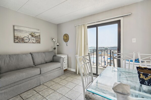 A living room with a grey sofa, a glass-top dining table, and a view of the city through a large window. - Beachfront Fourth-Floor Condo w/ Community Pool! (North Wildwood)