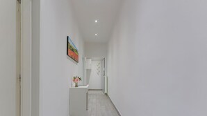Un pasillo estrecho con pared blanca, un cuadro, una pequeña barra blanca con un jarrón de flores y una puerta al final.
