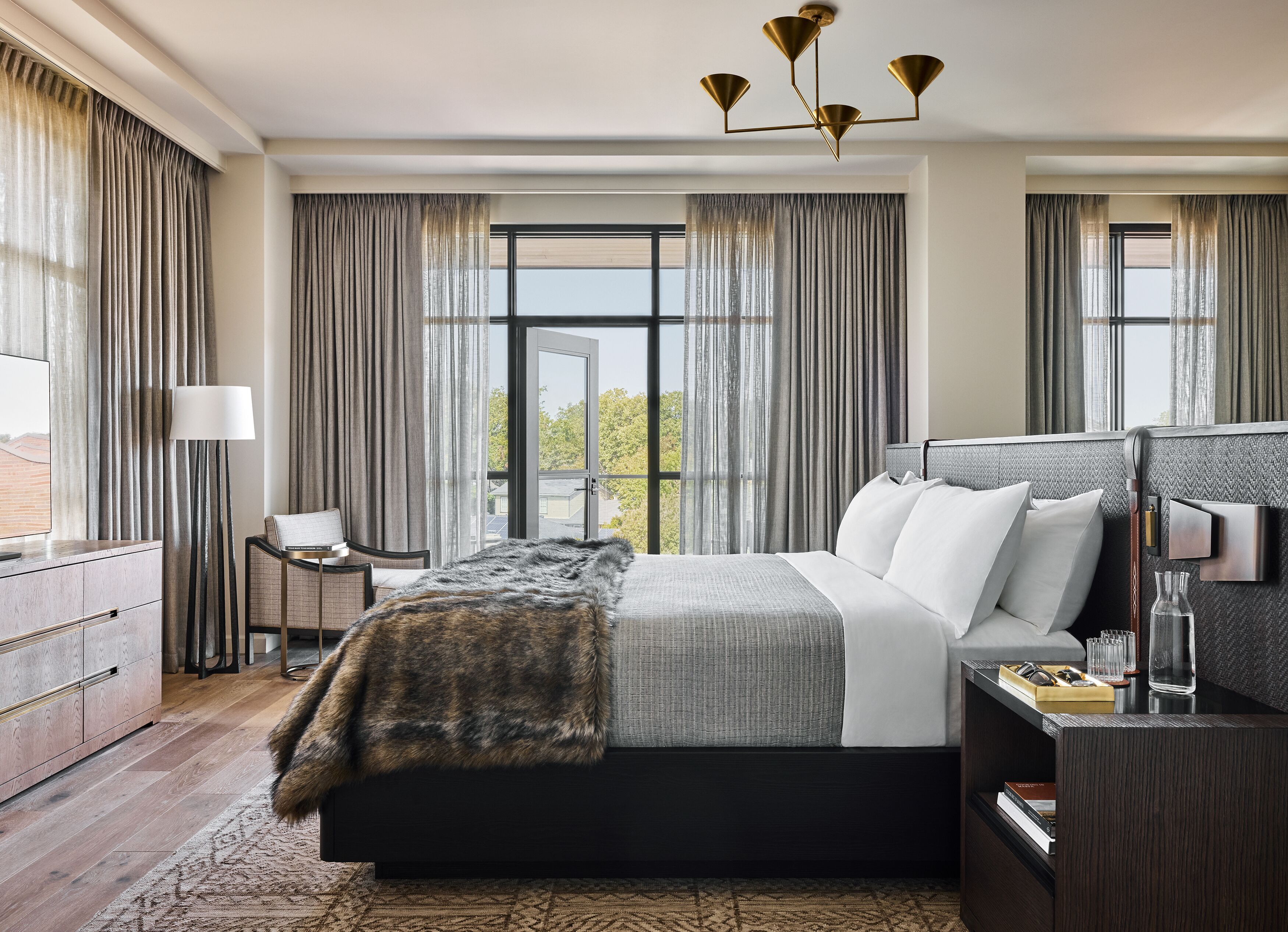 A modern hotel room with a large bed, a desk, a chair, and a view of the outdoors through large windows.