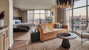 A modern hotel room with a large bed, a sofa, a dining table, and a view of the city through large windows. - Bowie House, Auberge Collection (Fort Worth)