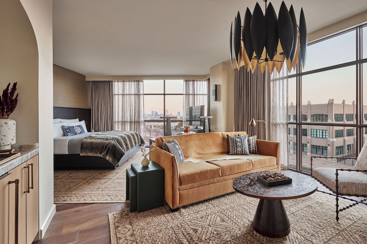 A modern hotel room with a large bed, a sofa, a dining table, and a view of the city through large windows.