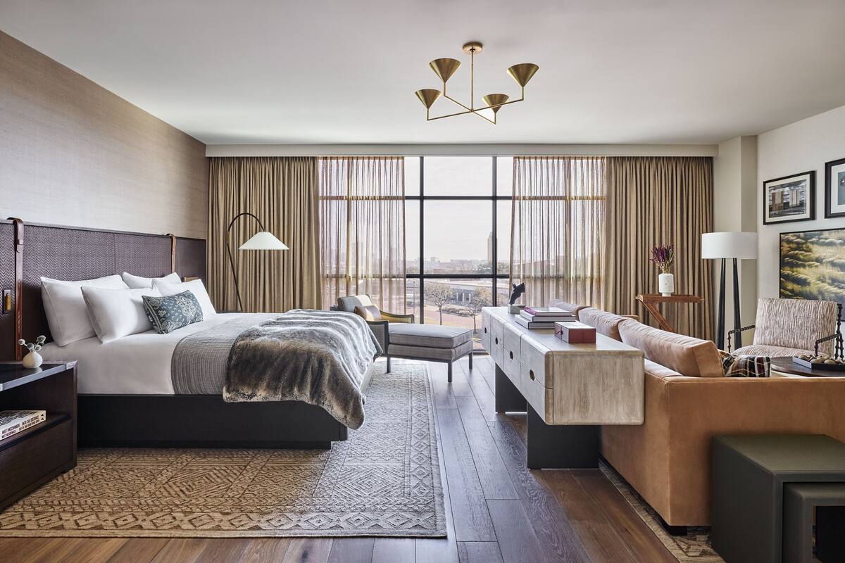 A modern hotel room with a large bed, a sofa, a desk, and a view of the city through floor-to-ceiling windows.