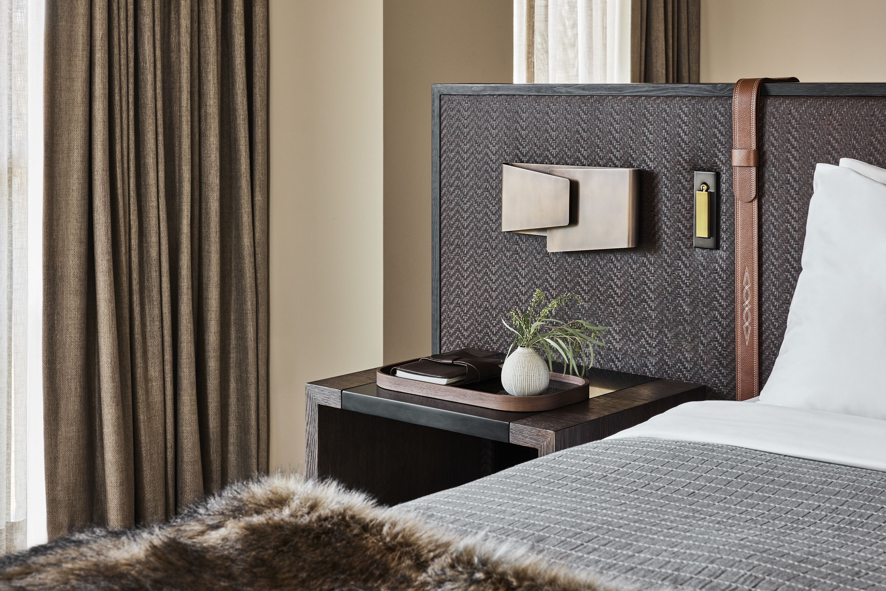 A modern hotel room with a large bed, a nightstand with a lamp, a small potted plant, and a view of the outside through curtains.