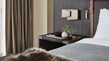 A modern hotel room with a large bed, a nightstand with a lamp, a small potted plant, and a view of the outside through curtains.