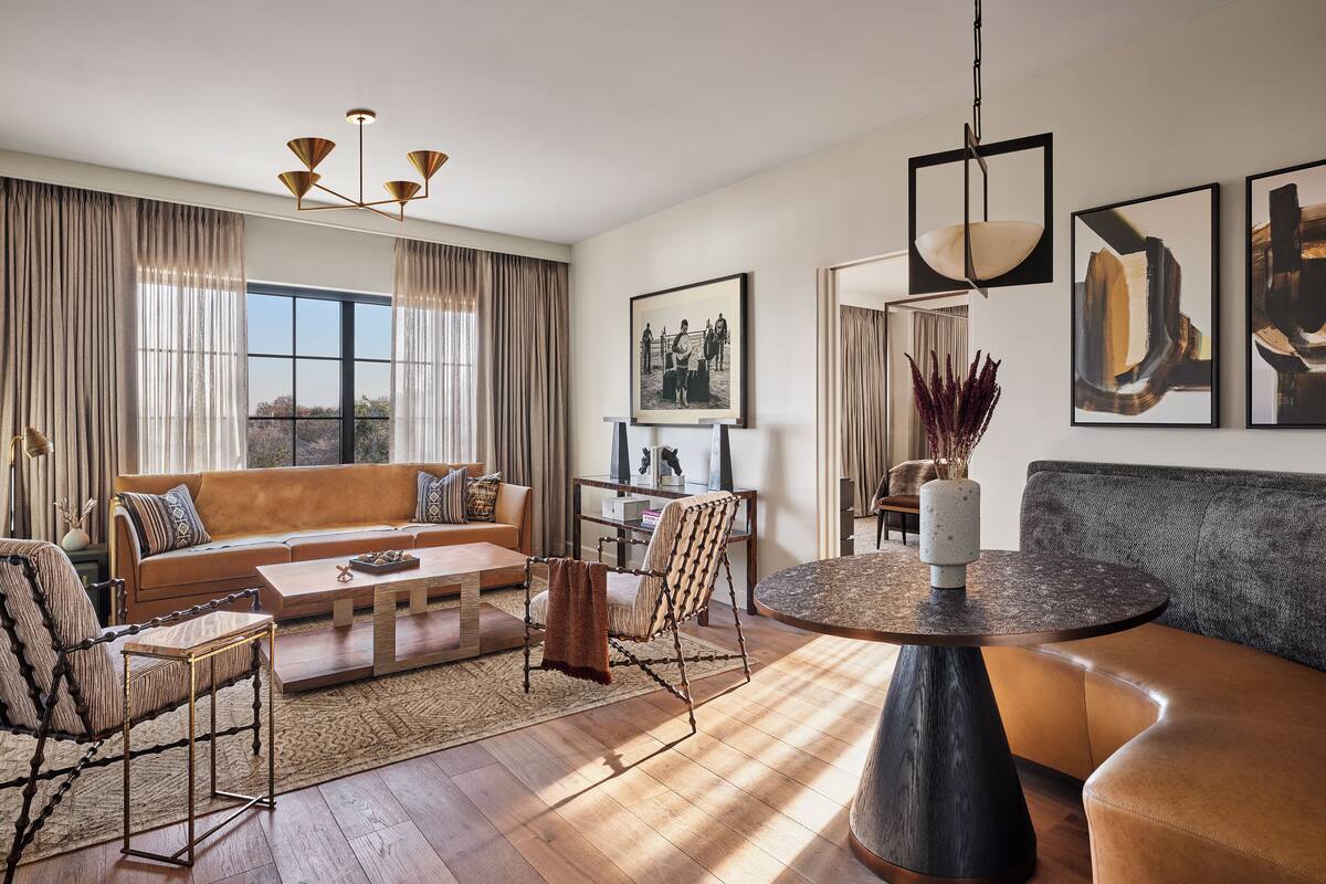A modern living room with a sofa, armchairs, a coffee table, and a dining table. There are curtains, a chandelier, and artwork on the walls.
