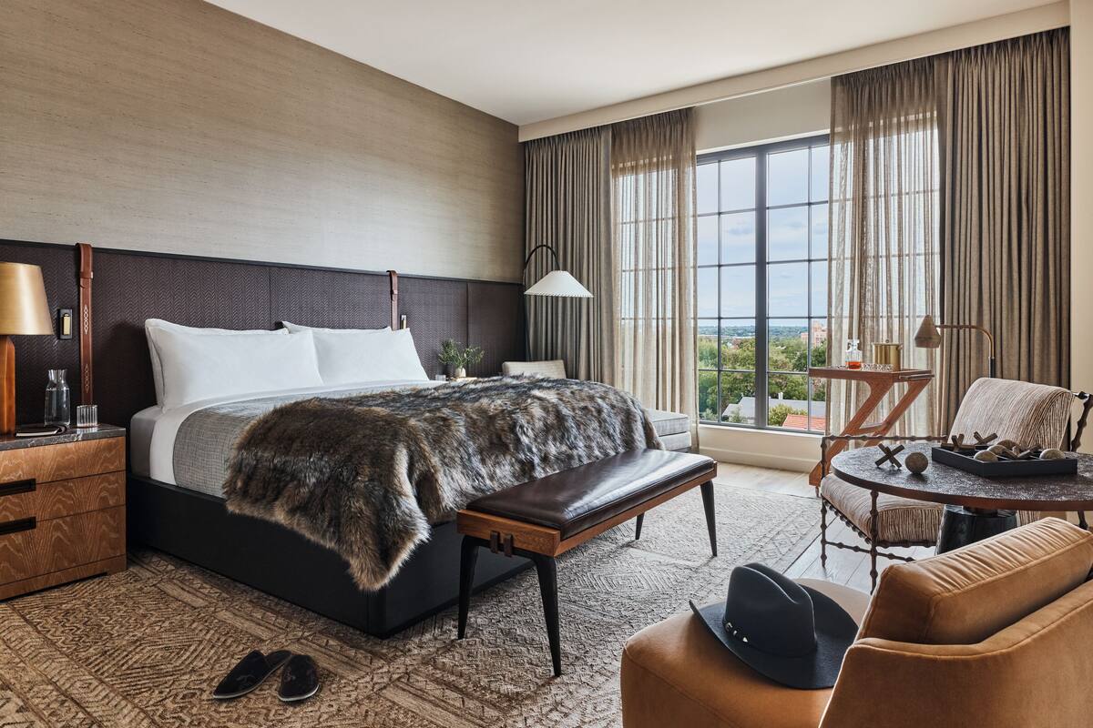 A modern hotel room with a large bed, a bench, a small table with a lamp, and a view of the city through large windows.