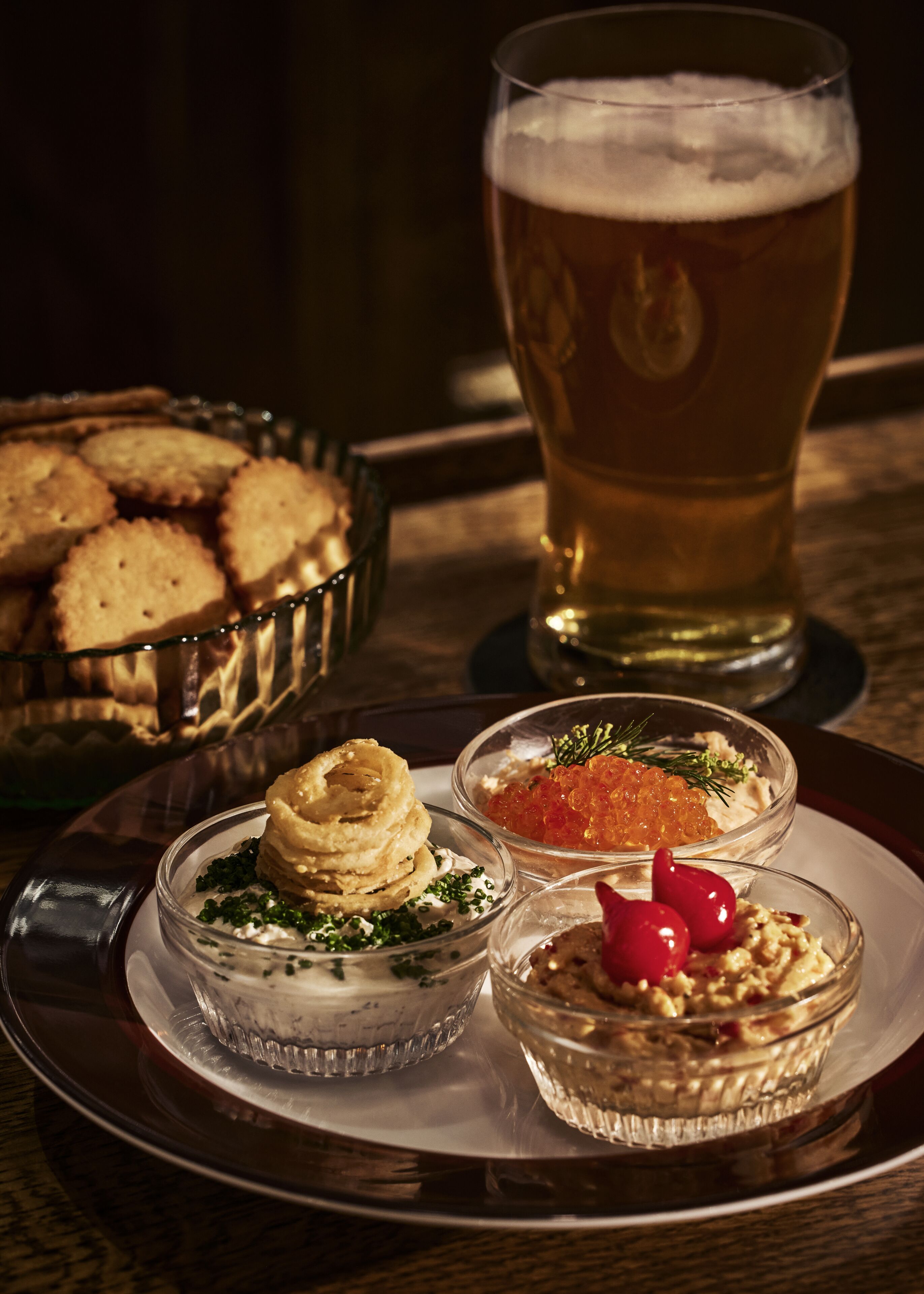 Una cerveza en un vaso, un plato con tres recipientes pequeños de comida y un bol de galletas.