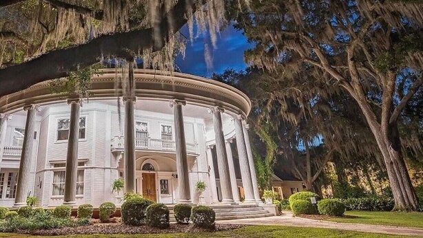Un grand bâtiment blanc de style classique, doté d’un portique circulaire central soutenu par des colonnes, et entouré d’une végétation luxuriante et d’arbres couverts de mousse pendant.