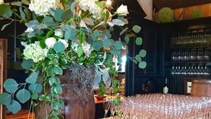 Un coin bar avec un comptoir en bois, un grand vase rempli de fleurs blanches et de feuillage vert, et une rangée de verres vides.