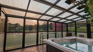 A hot tub in a covered outdoor area with large windows offering views of a garden and trees. - Panoramic Catskills Farmhouse and Spa (Harpersfield)