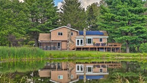 A two-story house with a deck, a pond in front, and a forested backdrop. - Panoramic Catskills Farmhouse and Spa (Harpersfield)