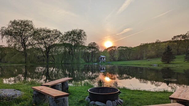 A serene lakeside setting with wooden benches and a fire pit, surrounded by trees and a small wooden structure in the distance. - Panoramic Catskills Farmhouse and Spa (Harpersfield)